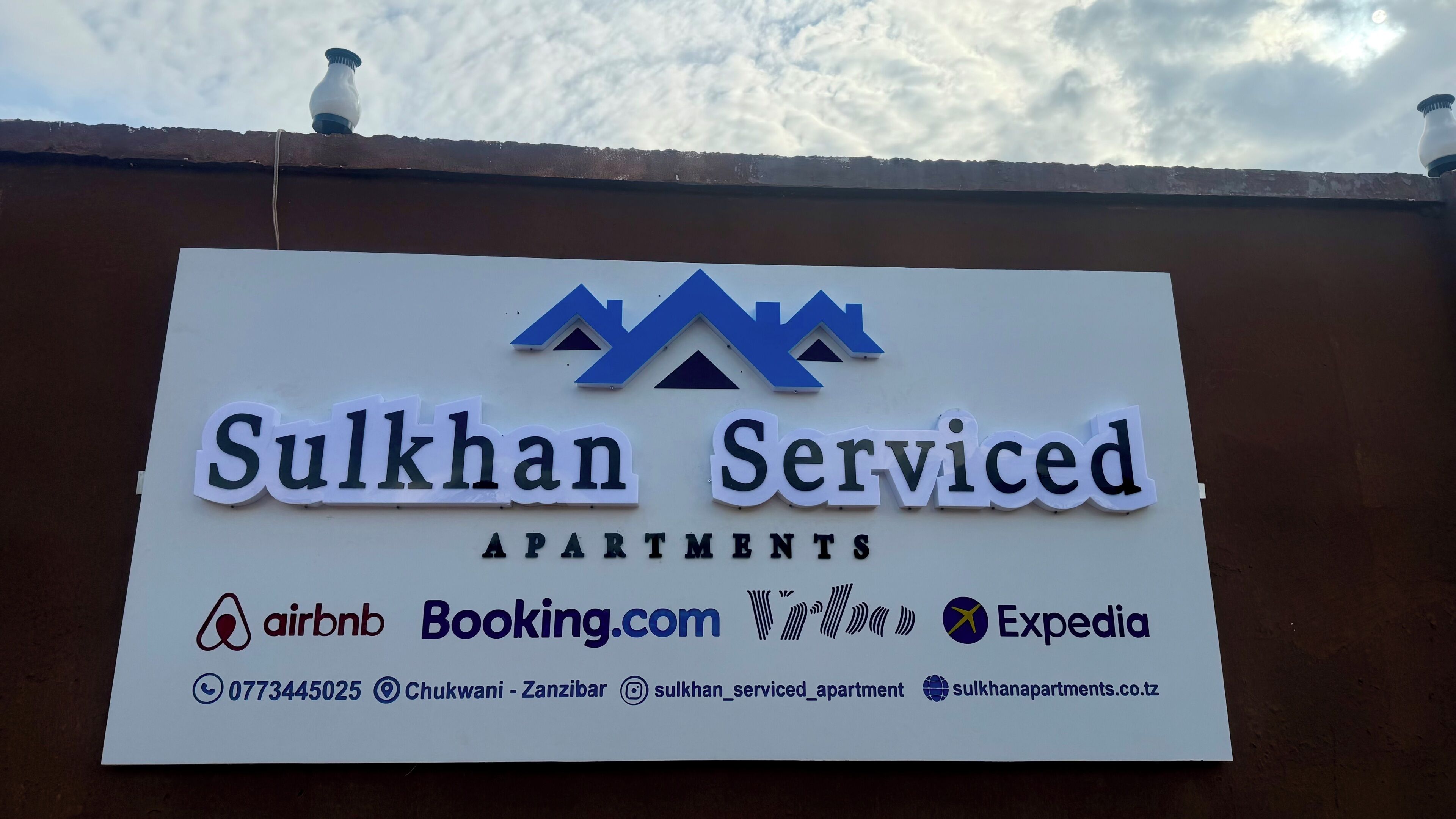 A sign for Sukhman Serviced Apartments with logos for Airbnb, Booking.com, and Expedia, and contact information.