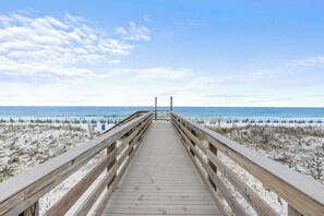 Ein hölzerner Steg führt zu einem Strand mit Dünen und dem Meer im Hintergrund.