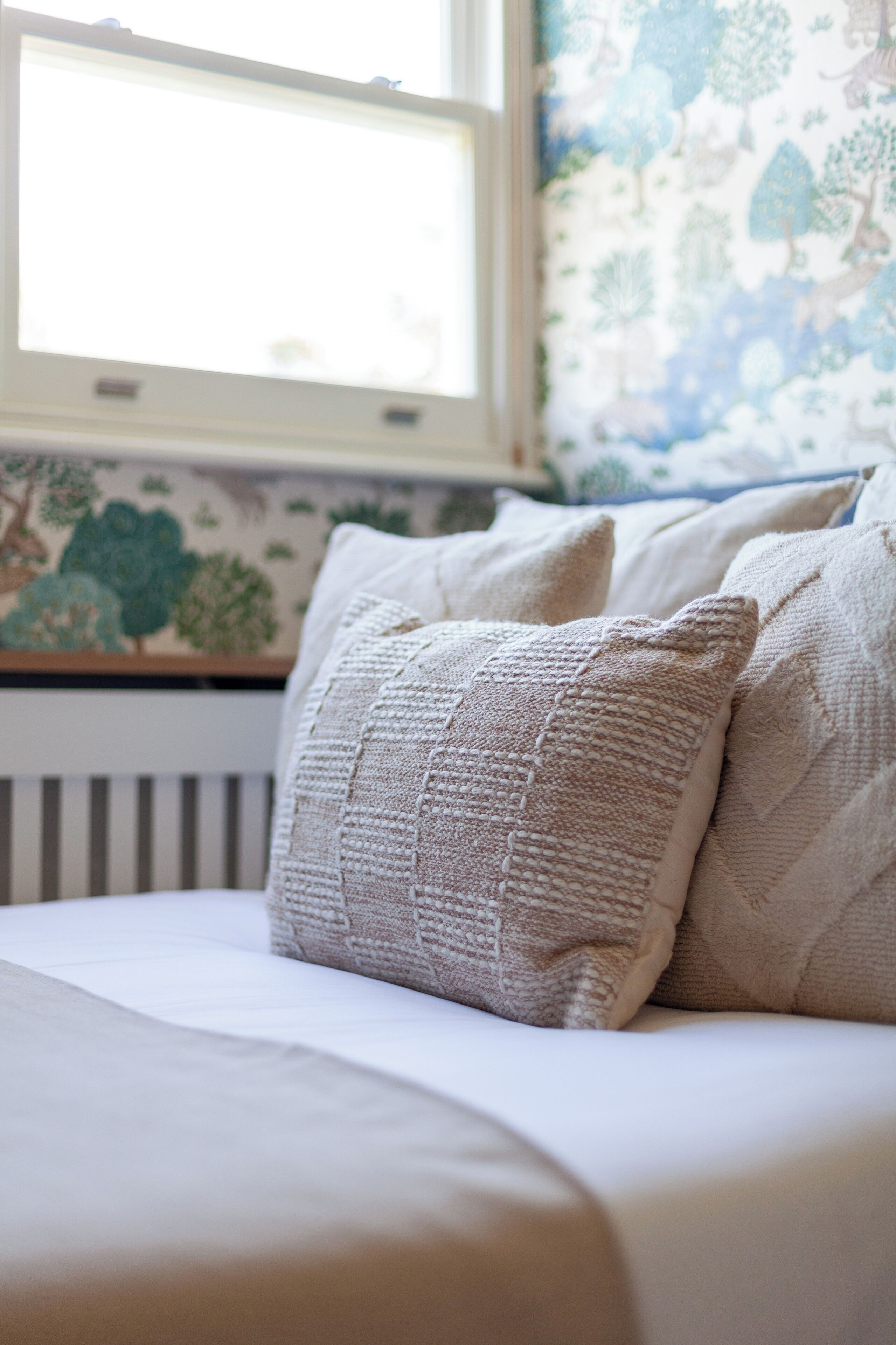 A bed with beige bedding and pillows, a window with a view of trees, and a radiator.