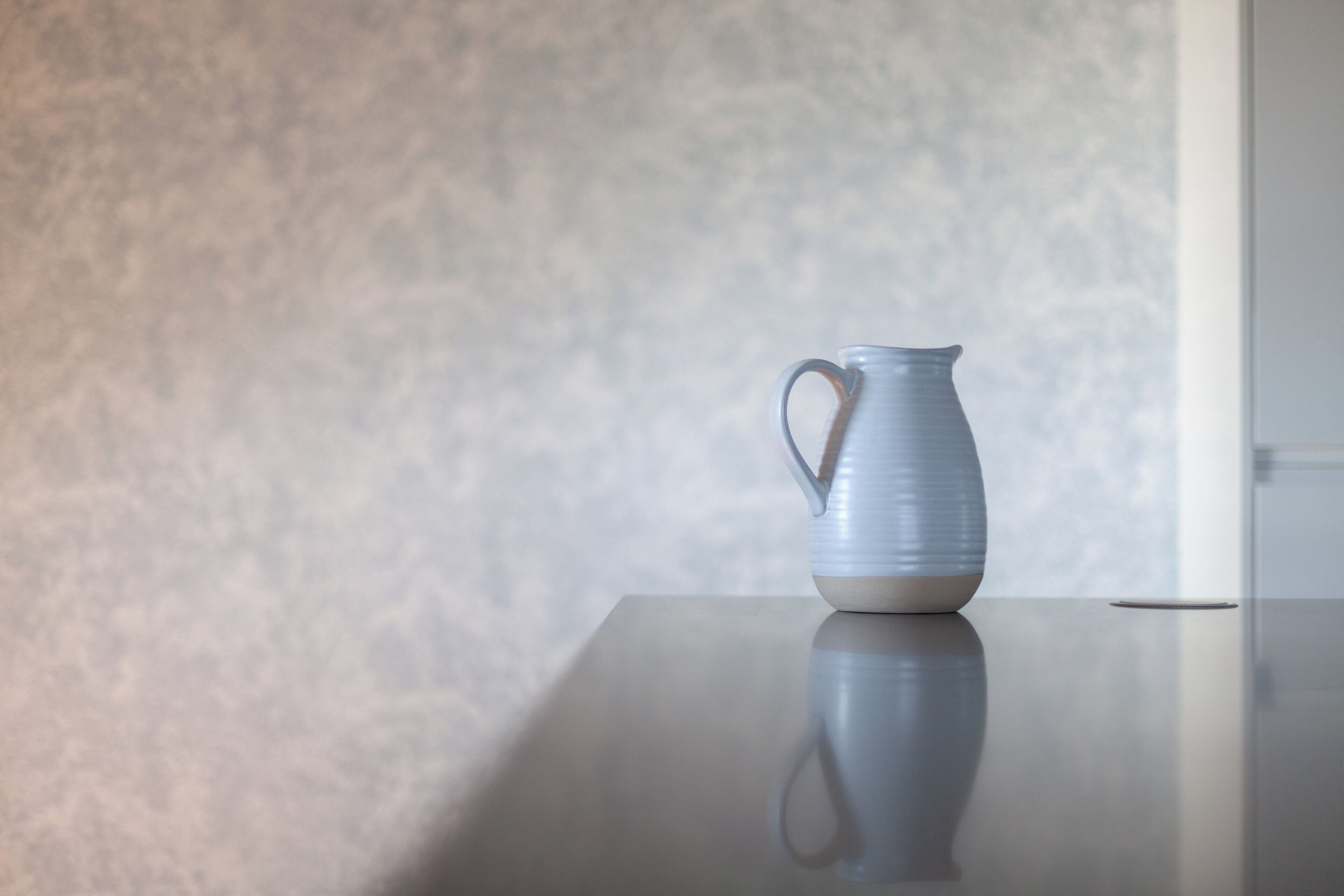 A white ceramic pitcher on a reflective surface against a textured wall.