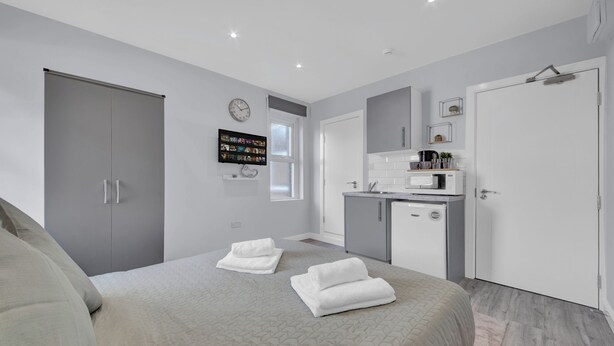 A modern bedroom with a bed, two white towels, a grey wardrobe, a small kitchenette, and a TV mounted on the wall. - Captivating 1-bed Studio in West Drayton (West Drayton)