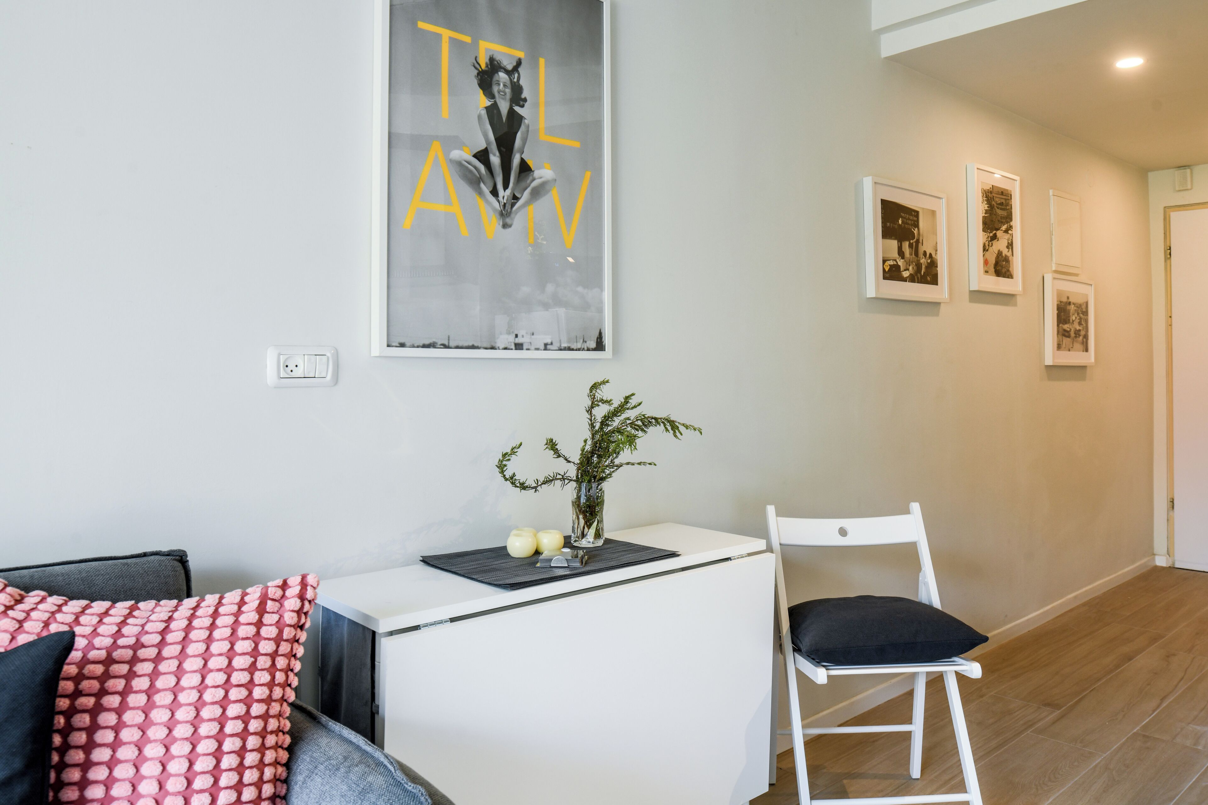 A modern living room with a white sofa, a small white table with a plant, and a white chair. There are framed pictures on the wall and a large poster above the table.