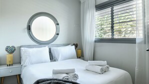 Een netjes opgemaakt bed met witte lakens en opgerolde handdoeken, een ronde spiegel aan de muur en een raam met zonwering.