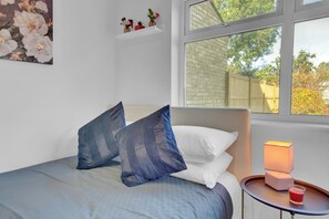 A bedroom with a bed, pillows, a bedside table with a lamp, and a window with a view of a brick wall and trees. - Charming Serviced Studio West Drayton London UB7 (West Drayton)