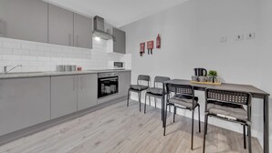 Una cocina moderna con armarios grises, una encimera blanca y un horno empotrado. Hay dos sillas con cojines grises y una mesita con una planta.
