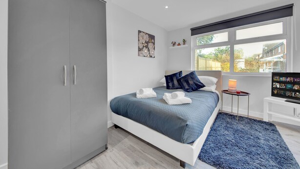 Un dormitorio moderno con un armario grande gris, una cama con colcha azul, almohadas blancas y una alfombra azul. Hay una mesita con una lámpara y un televisor sobre un mueble.