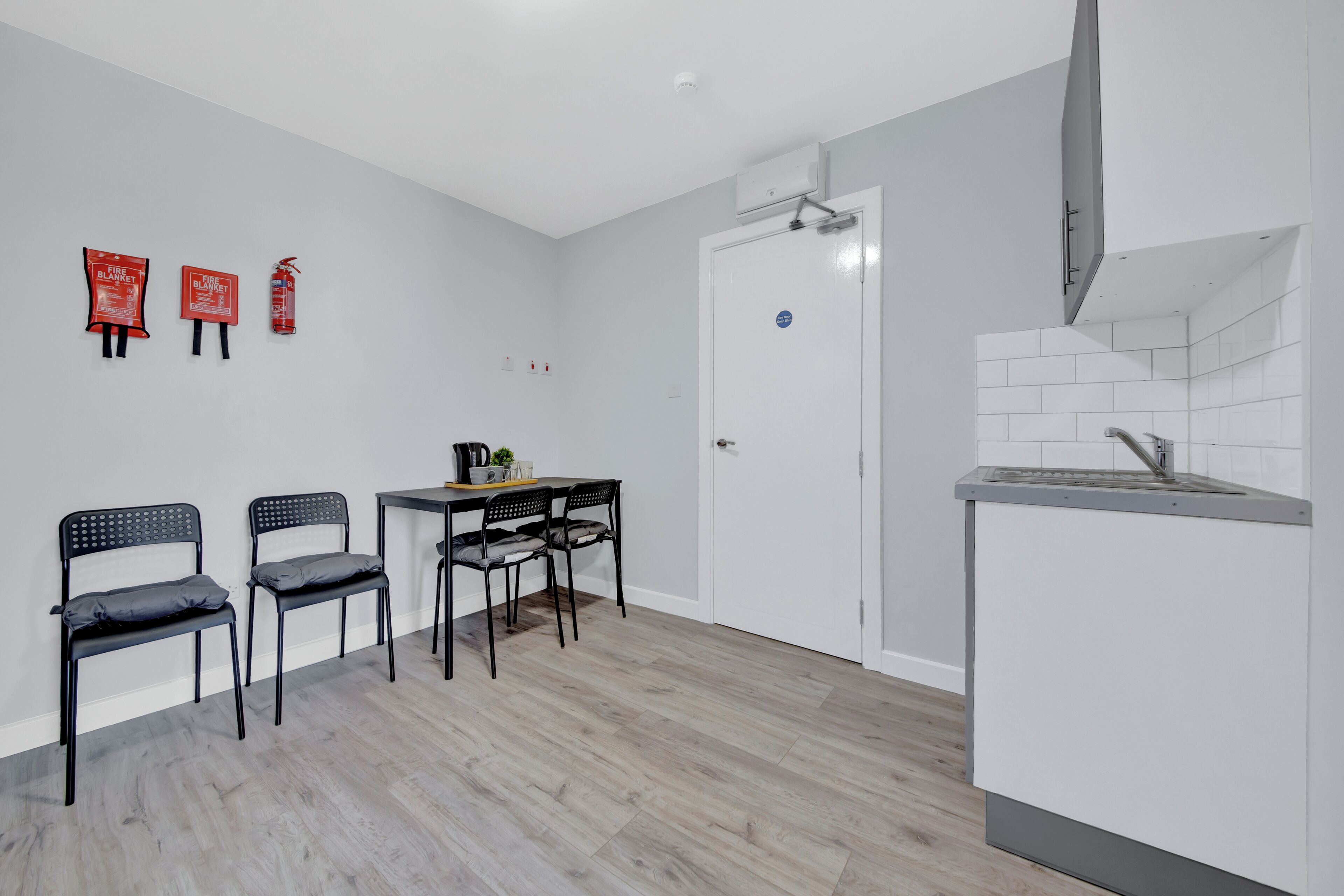 A modern kitchen with a white countertop, a built-in oven, and a sink. There are two black chairs and a small table with a kettle and a cup.