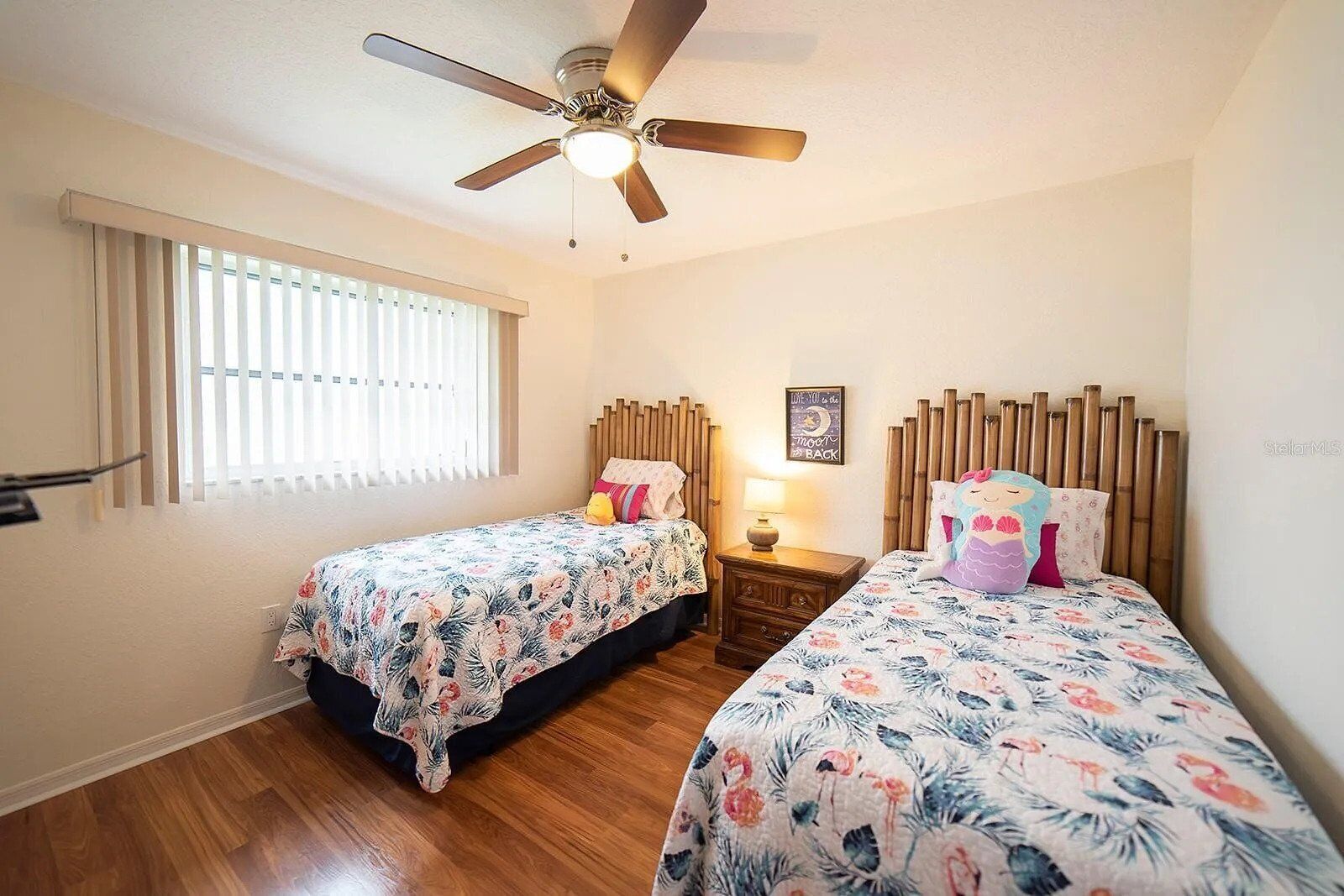 A bedroom with two single beds, a ceiling fan, a window with blinds, and a nightstand with a lamp.