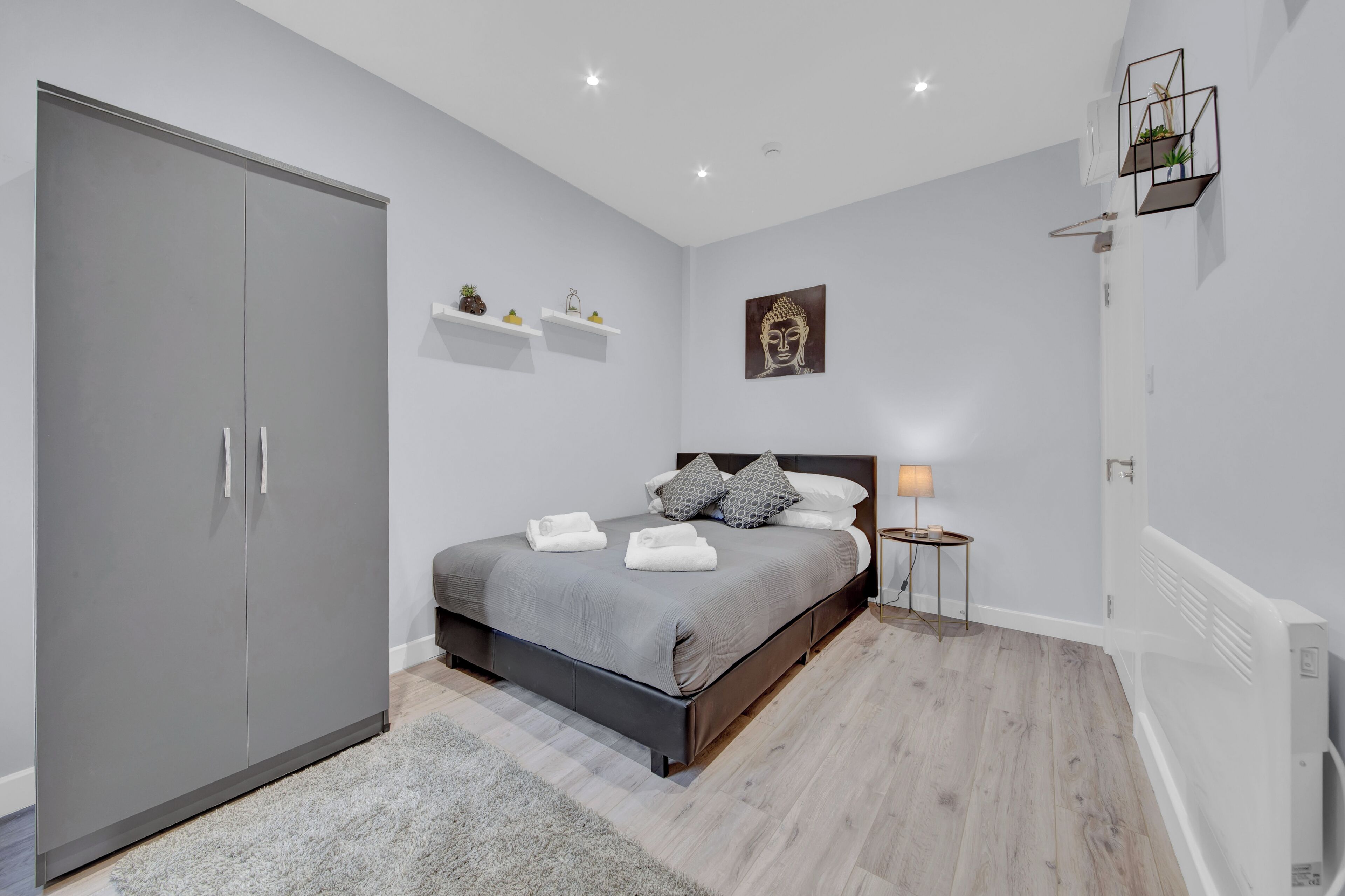 A modern bedroom with a grey wardrobe, a bed with grey bedding, a bedside table with a lamp, and a wall-mounted shelf with decorative items.