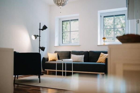 A modern living room with a dark-colored sofa, a black chair, and a floor lamp. There are two windows with white frames and a view of greenery outside. - Appartement Residenz Rosenbach - Gemütliche Unterkunft im Herzen von Würzburg (Würzburg)