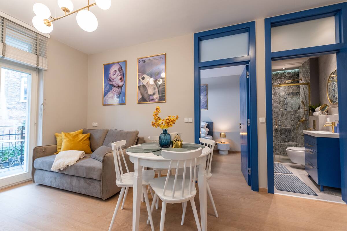 A modern living room with a grey sofa, white chairs, and a dining table with a vase of flowers. There is a blue-framed door leading to a bathroom with a white sink and a mirror.