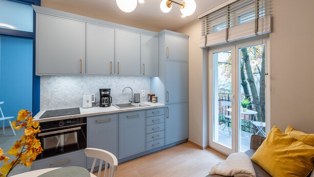 A modern kitchen with grey cabinets, a built-in oven, and a coffee maker. There is a dining area with a table and chairs, and a window with blinds. - City Lights Mostowa (Kraków)