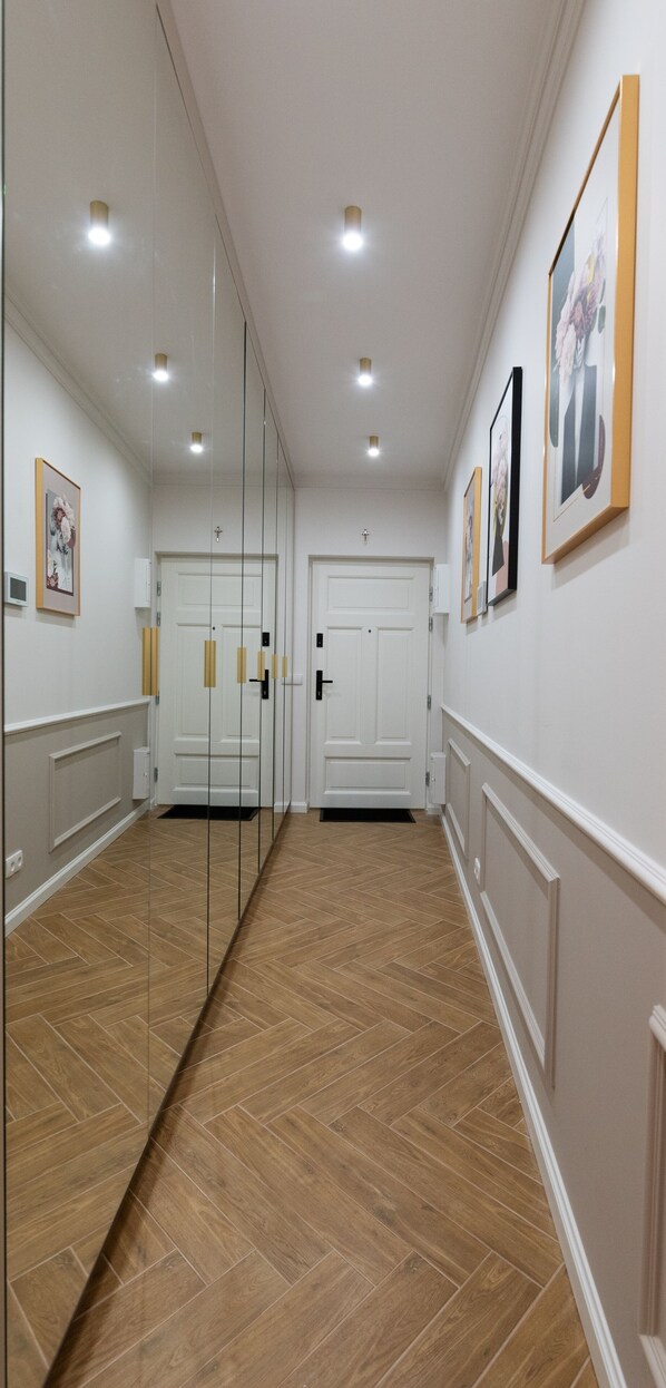 A hallway with wooden flooring, white walls, and a mirrored wall. There are framed pictures on the wall and a door at the end.