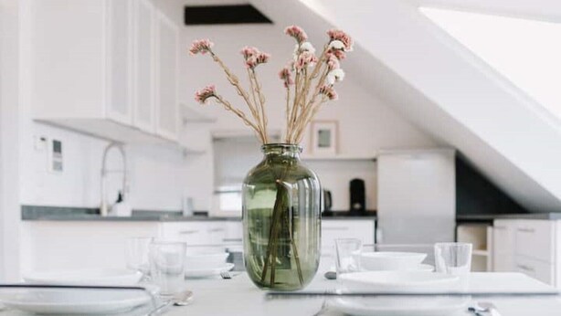 Eine moderne Küche mit weißen Schränken, einem Esstisch für zwei Personen und einer Vase mit Trockenblumen auf dem Tisch.