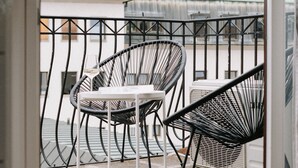 Ein Balkon mit zwei schwarzen Drahtstühlen und einem kleinen Tisch, mit Blick auf eine Stadtansicht.