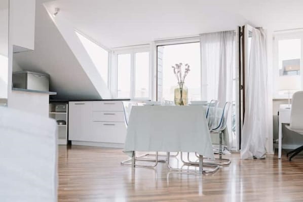 Ein moderner, hell erleuchteter Essbereich mit Tisch für zwei Personen, einem weißen Schrank und einem großen Fenster mit Vorhängen.