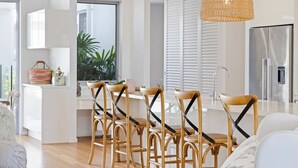 A modern kitchen with white cabinetry, a stainless steel refrigerator, and a dining area with wooden chairs and a woven pendant light. - Hampton Beach House on the Waterfront (Buddina)