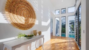 A modern interior with a woven wall art piece, a white console table with decorative items, and a view through large glass doors to an outdoor area. - Hampton Beach House on the Waterfront (Buddina)
