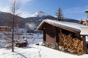 Una cabaña de madera con techo nevado, una pila de leña y montañas al fondo.
