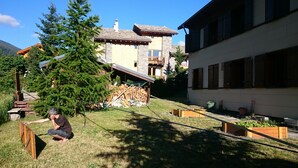 Zona residencial con terraza de madera, un gran árbol de hoja perenne y un edificio de piedra con techo inclinado.