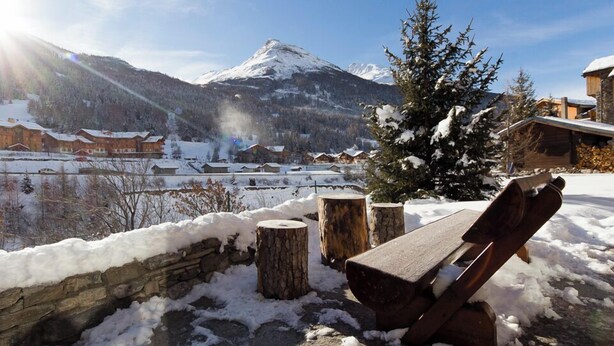 Un banco de madera en un espacio exterior nevado con un muro de piedra y troncos de madera, con vistas a un pueblo con edificios cubiertos de nieve y una montaña al fondo.