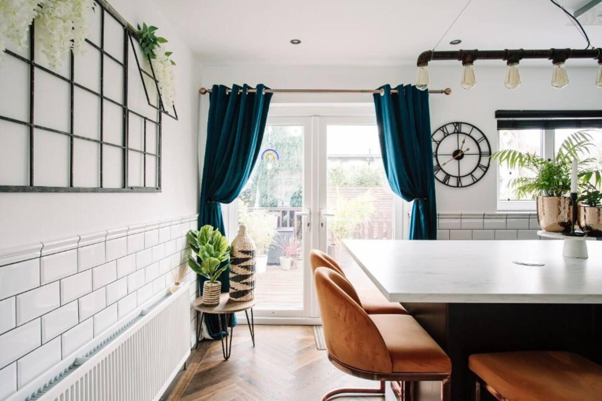 A modern kitchen with a central island, blue curtains, a clock, and potted plants.