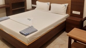 A single bed with white bedding, two pillows, and a striped pillow. There is a wooden nightstand with a lamp and a wooden wardrobe with two doors. - Rani Residency (Nagercoil)