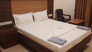 A neatly made bed with white linens and two striped pillows, a wooden headboard, a small wooden nightstand, a chair, and a table with a brown tablecloth. - Rani Residency (Nagercoil)