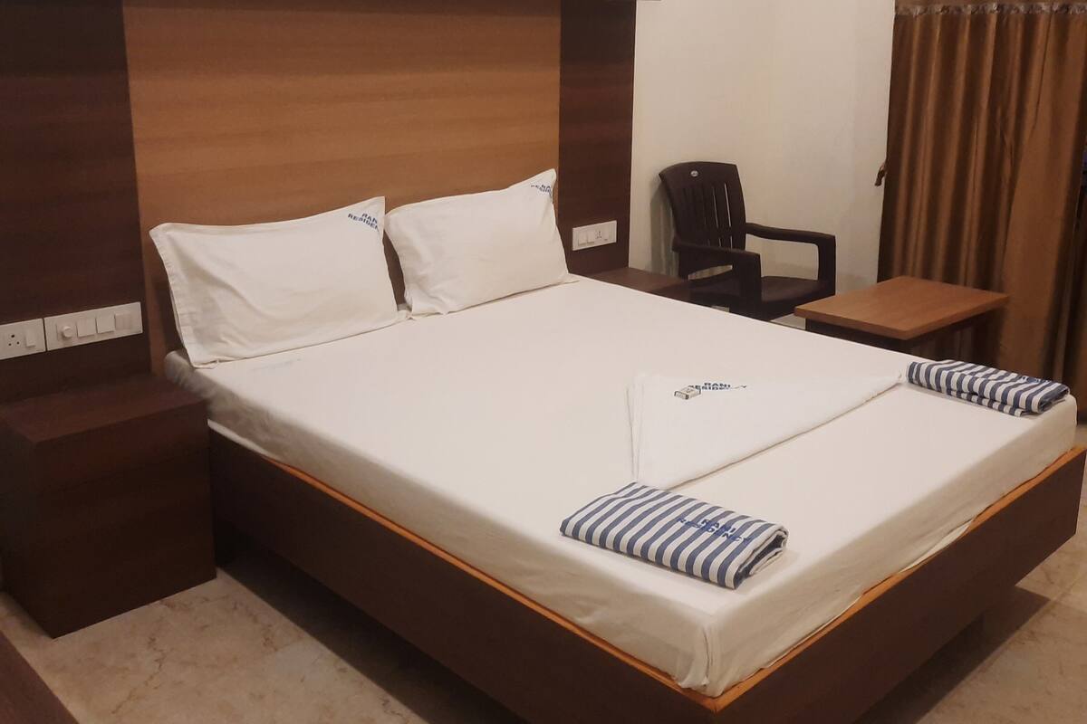 A neatly made bed with white linens and two striped pillows, a wooden headboard, a small wooden nightstand, a chair, and a table with a brown tablecloth.