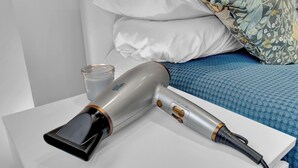 A hair dryer on a white bedside table with a glass of water beside it, a blue bedspread, and white pillows in a hotel room.