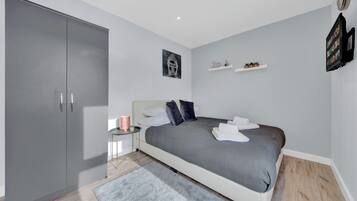 A modern bedroom with a large grey wardrobe, a neatly made bed with a grey blanket, a small bedside table with a lamp, and a wall-mounted shelf with decorative items.