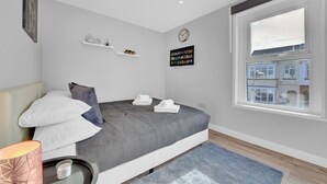 A modern bedroom with a bed, pillows, a bedside table with a lamp, a clock, and a window with a view of buildings.