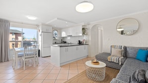 A modern kitchen with white cabinets, a dining area with a white table and chairs, a grey sofa with patterned cushions, and a round coffee table. - Belle Escapes - Sunny Siesta at Glenelg (Glenelg)