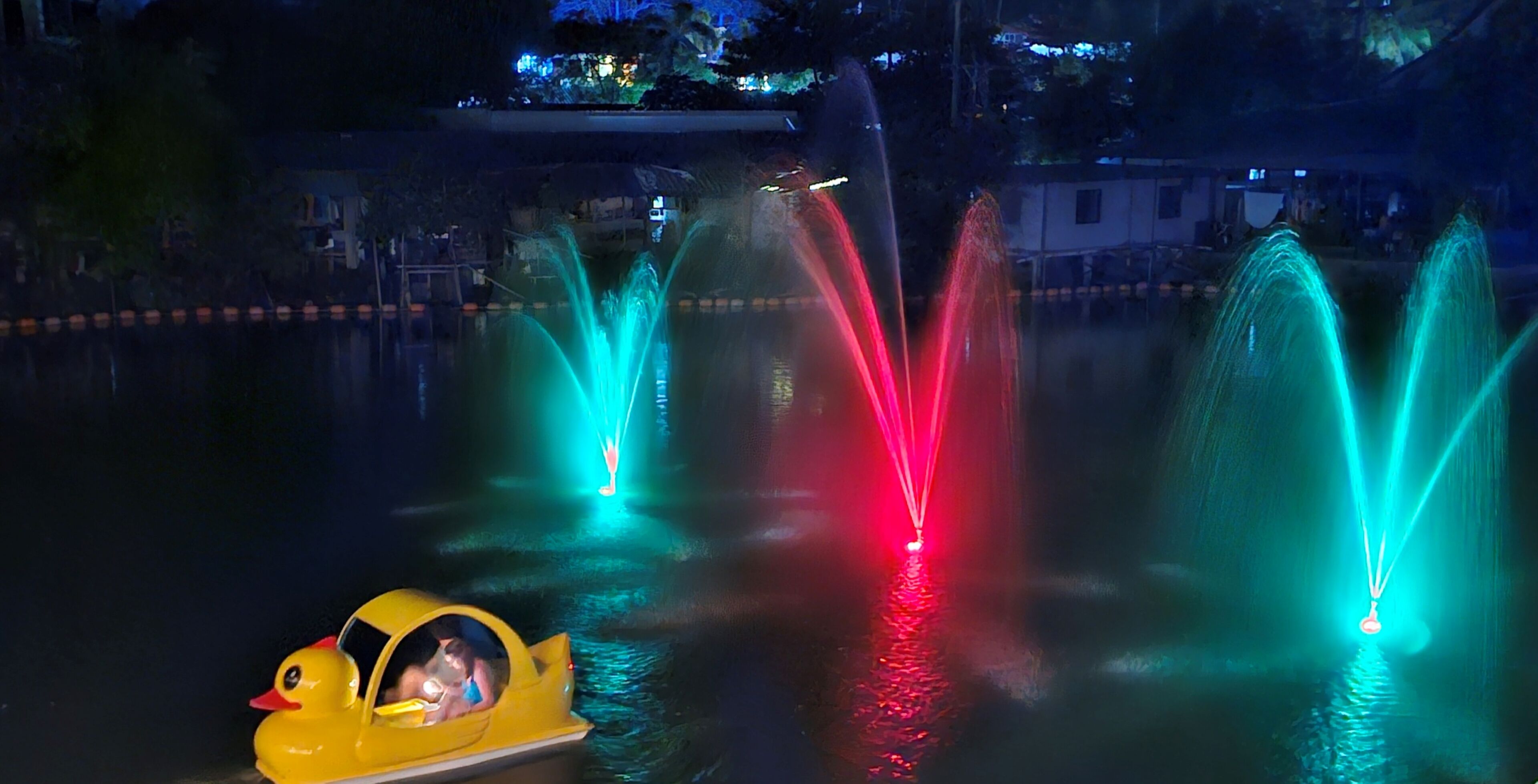 A nighttime view of a water fountain with colorful lights, a duck-shaped boat, and a building in the background.