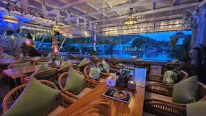 A restaurant with wicker furniture, a wooden table with a menu, and a view of a water body and buildings. - Hideout Hostel (Ko Pha-ngan)