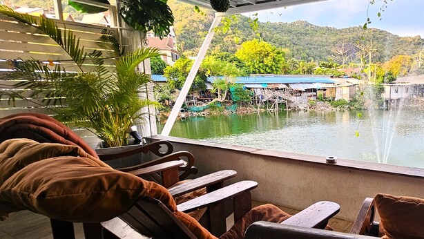 A restaurant with outdoor seating overlooking a body of water, featuring wooden tables and chairs with cushions. - Hideout Hostel (Ko Pha-ngan)