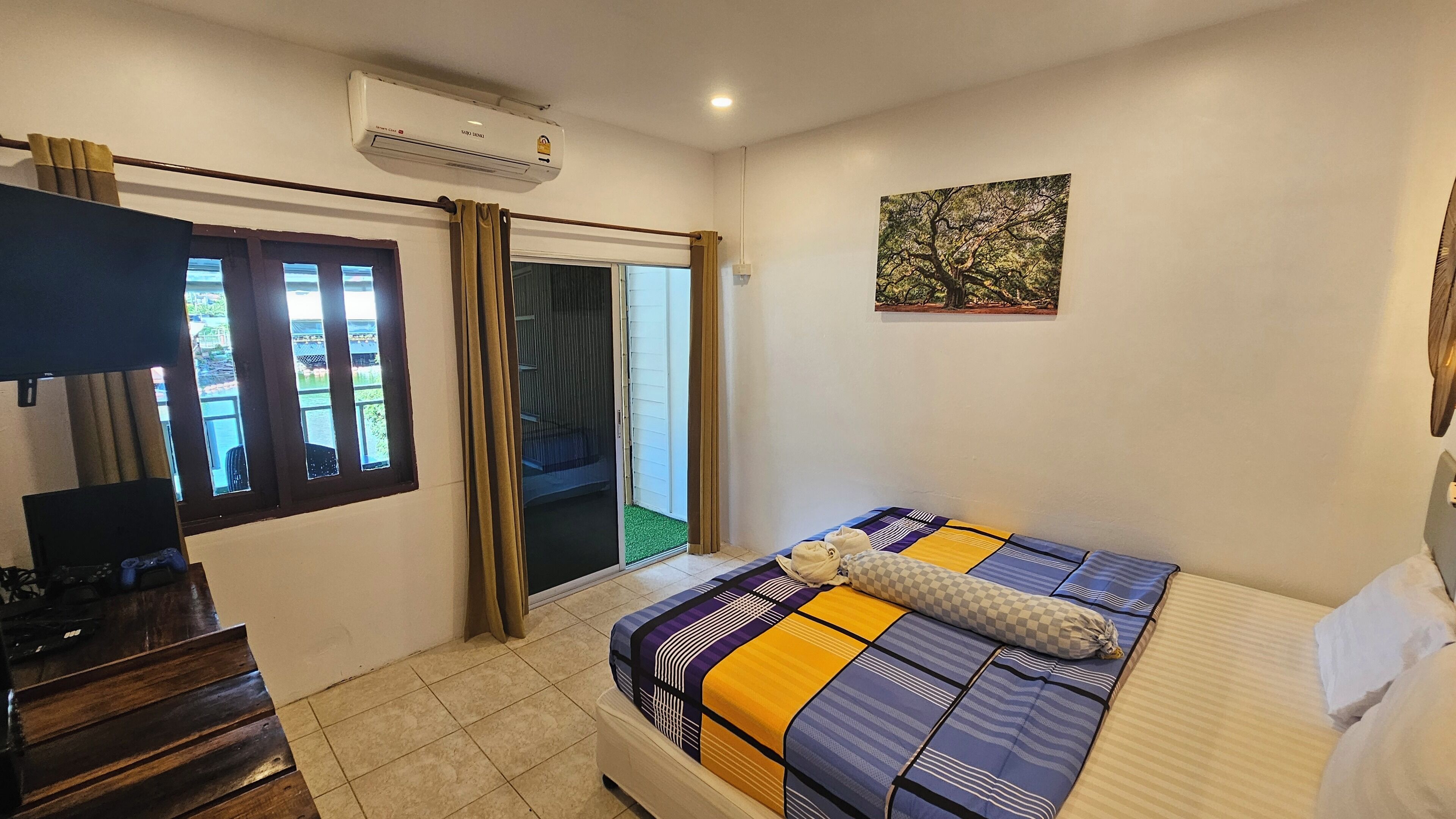 Un dormitorio con cama, escritorio, aire acondicionado, ventana con cortinas y puerta que da a un balcón.