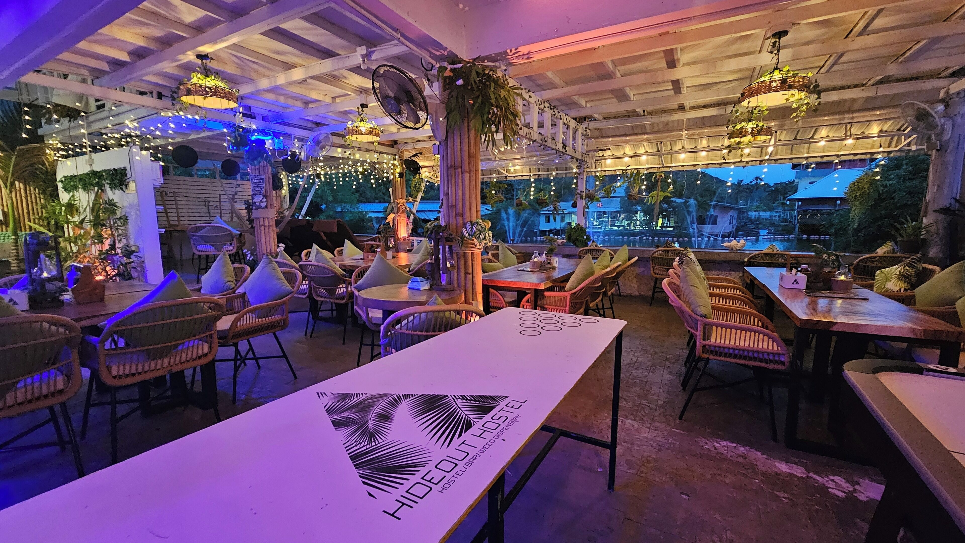 An indoor dining area with wicker chairs and tables, a view of the outside, and hanging plants.