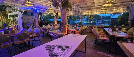 An indoor dining area with wicker chairs and tables, a view of the outside, and hanging plants.