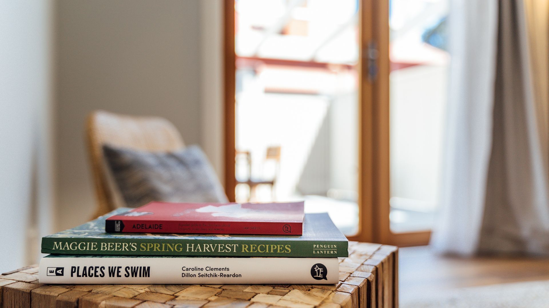 A stack of books on a wooden table in a well-lit room with a view of a window and a chair in the background.