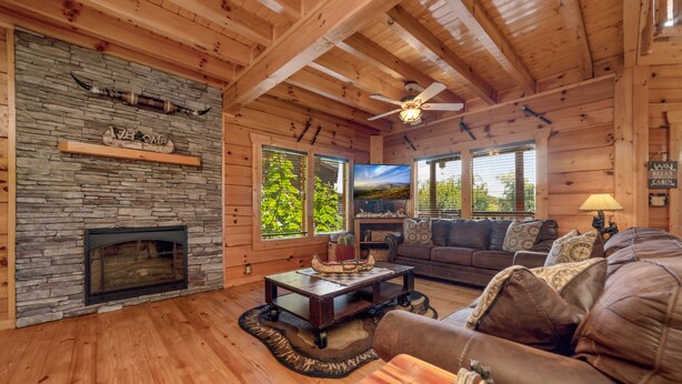 A log cabin interior with a stone fireplace, wooden floors, a ceiling fan, a living area with a sofa and armchairs, and large windows with a view of greenery.