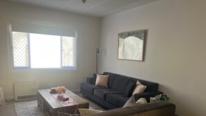 A living room with a dark blue sofa, a wooden coffee table with a vase of flowers, a large window with blinds, and a framed picture on the wall.