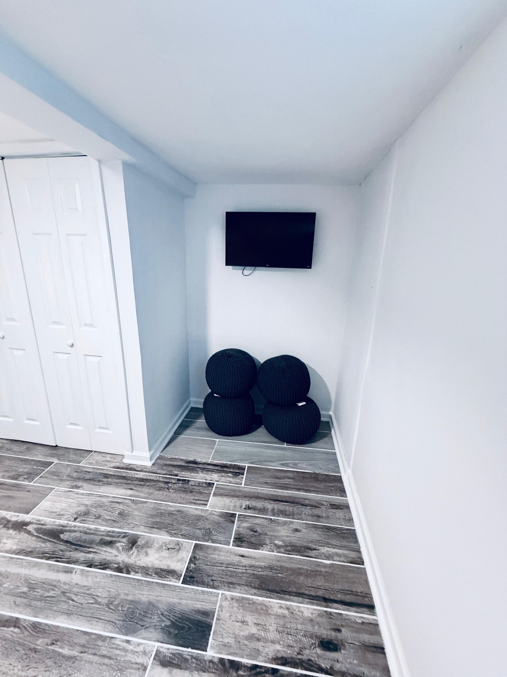 Een kamer met een witte deur, een wandmontage flatscreen televisie en vier ronde, zwarte poefjes tegen de muur gestapeld.