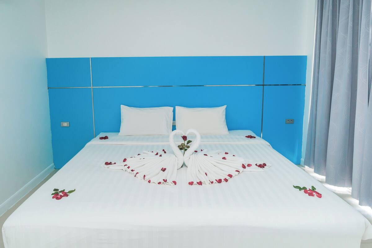 A hotel room with a blue headboard, white bedding, and a decorative swan made of rose petals and a rose on the bed.