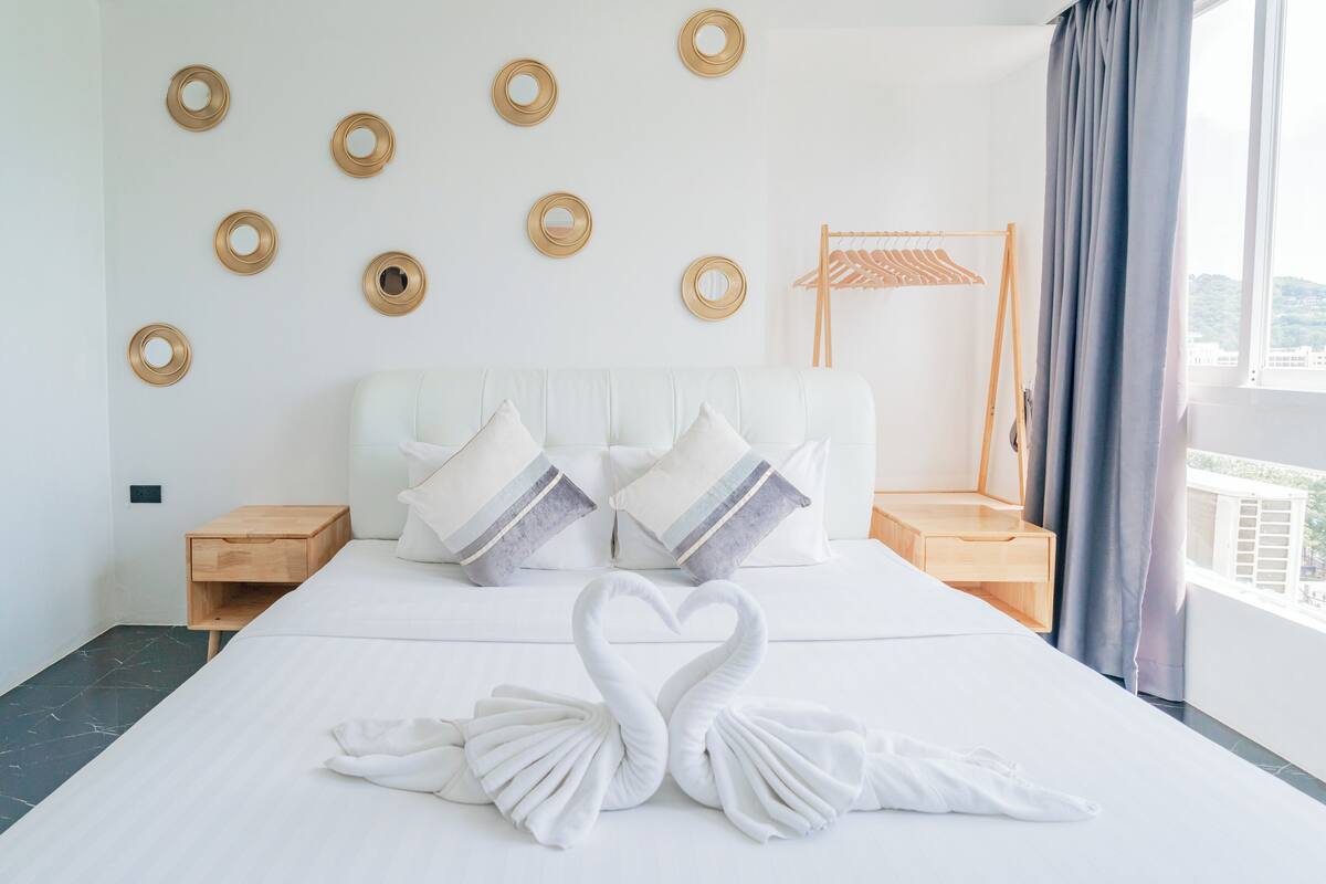 A hotel room with a neatly made bed, a wall-mounted circular mirror, a wooden nightstand, and a window with curtains.