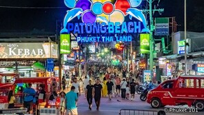 Uma rua movimentada com uma grande placa iluminada com a mensagem 'Welcome to Patong Beach, Phuket, Thailand'.