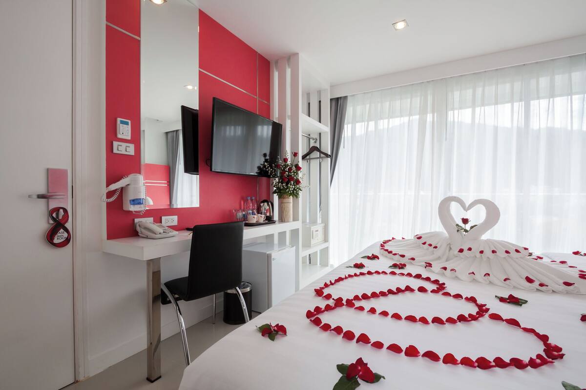 A hotel room with a bed adorned with rose petals and a heart-shaped decoration, a desk with a phone and a mirror, and a TV mounted on the wall.