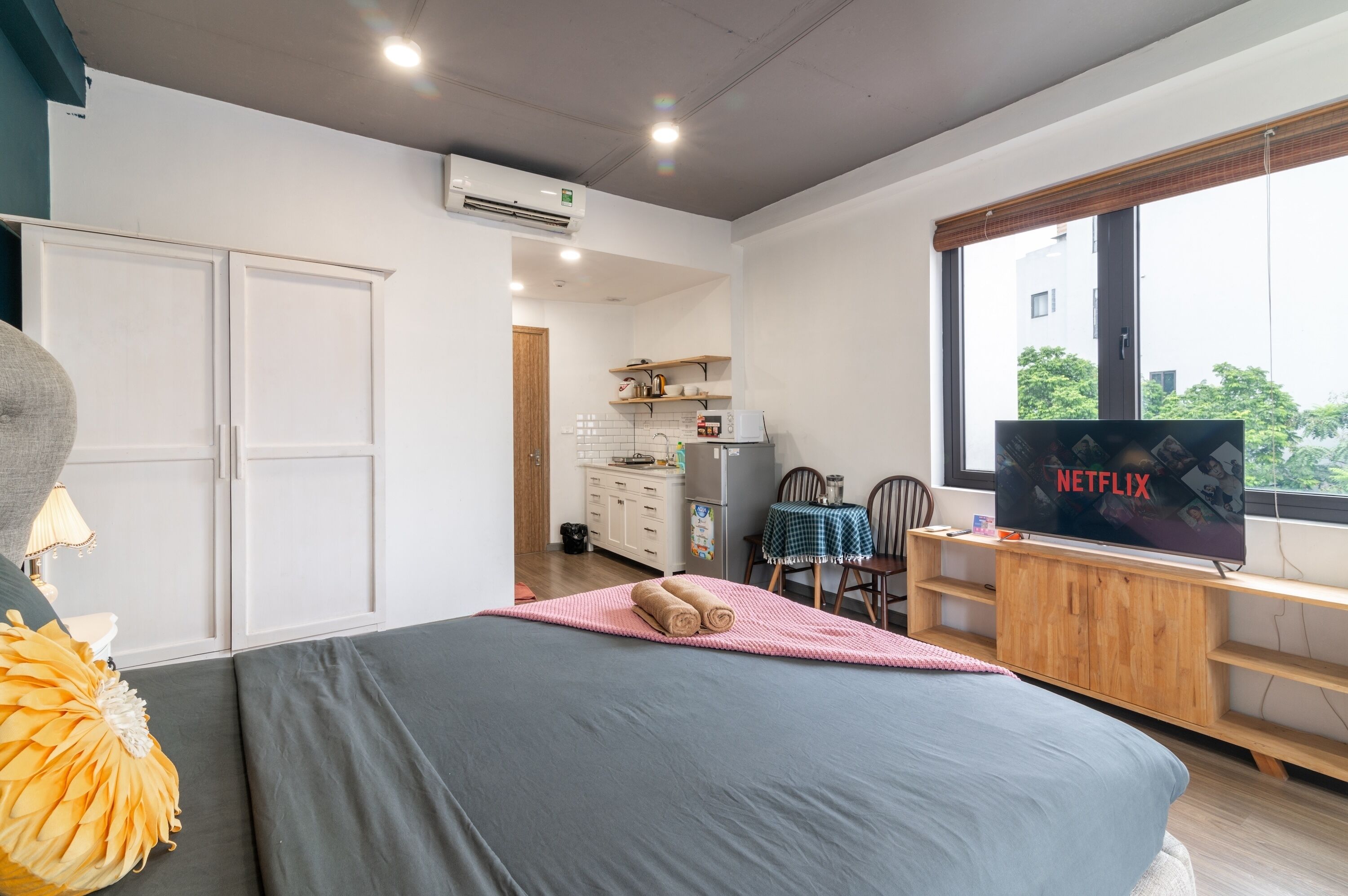 A modern bedroom with a large bed, a TV, and a kitchenette in the background.
