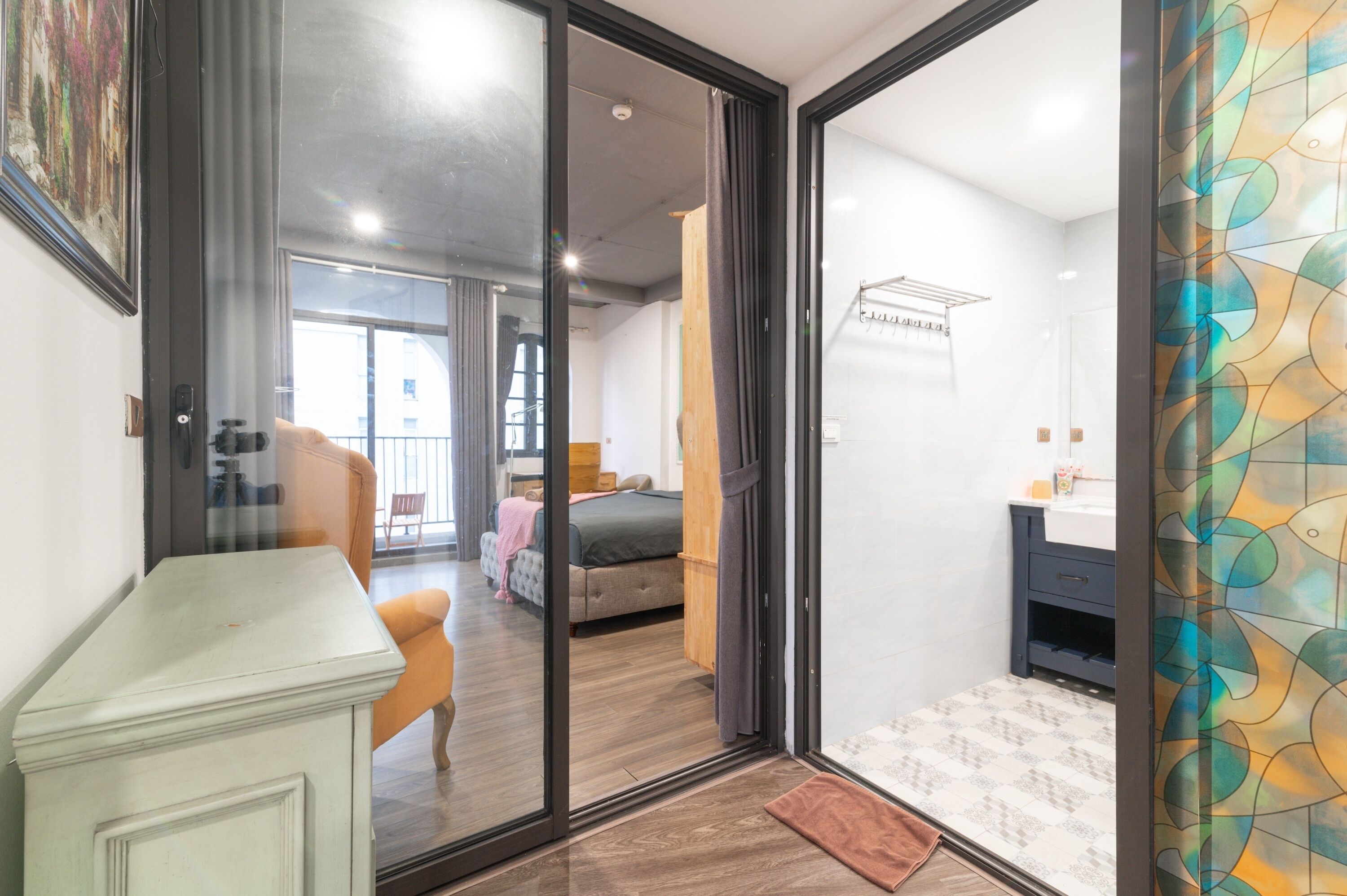 Una habitación de hotel moderna con una puerta de vidrio que da a un balcón, una cama con manta gris y rosa, un escritorio pequeño y un baño con espejo y toallero.