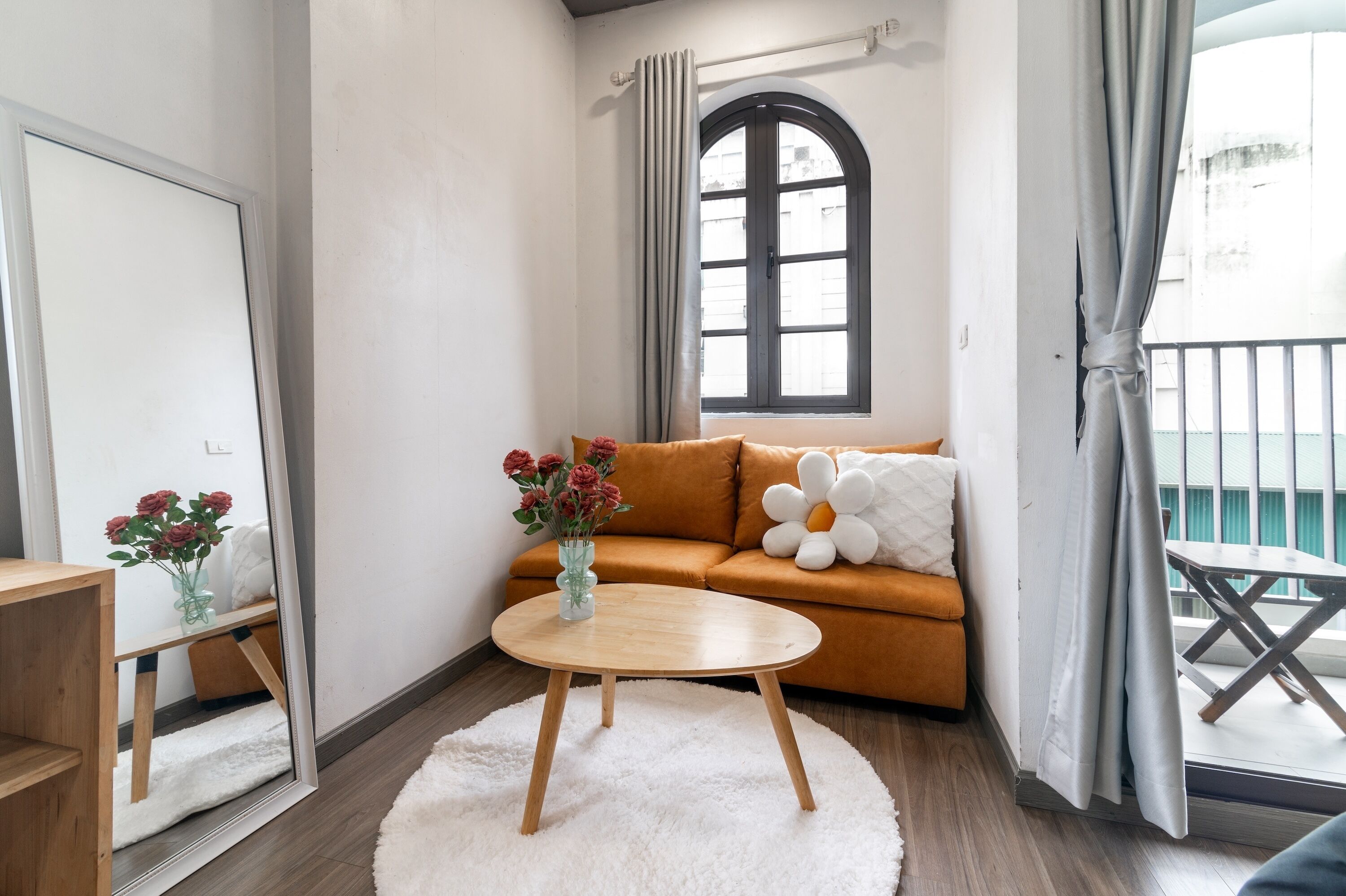 A cozy living room with a sofa, a round wooden table, a vase with flowers, and a large window with curtains.
